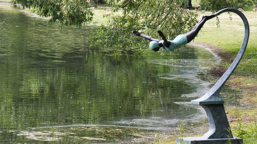 A sculpture of a person diving into a body of water positioned on the edge of the lake, with the figure in mid-dive, arms extended forward and legs together. The background includes lush greenery and trees, with their reflections visible on the calm surface of the water. The sculpture is made of metal and is mounted on a curved pedestal that supports it in its diving pose.